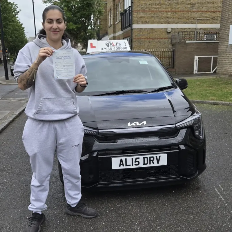 A new driver in a grey tracksuit holding up a pass certificate beside the black learner car