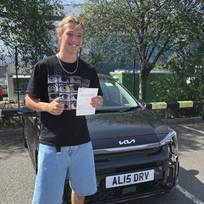 A new driver in a black T-shirt holding up a pass certificate in front of the learner car