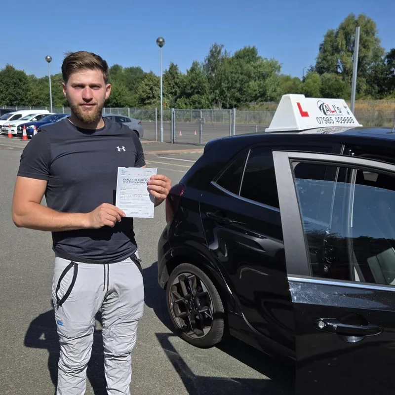 A new driver holding a pass certificate beside the learner car in a car park