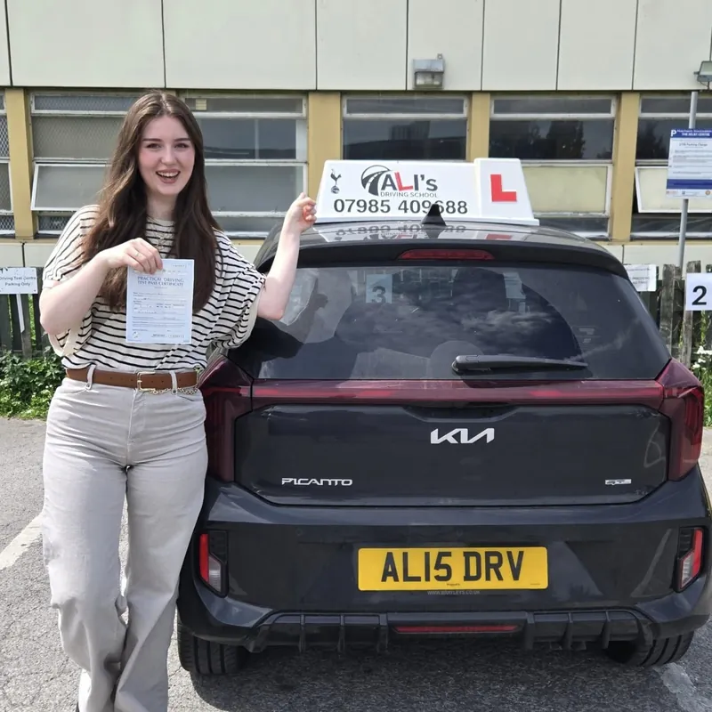 A new driver holding up a pass certificate behind the learner car, its roof sign showing Ali’s number