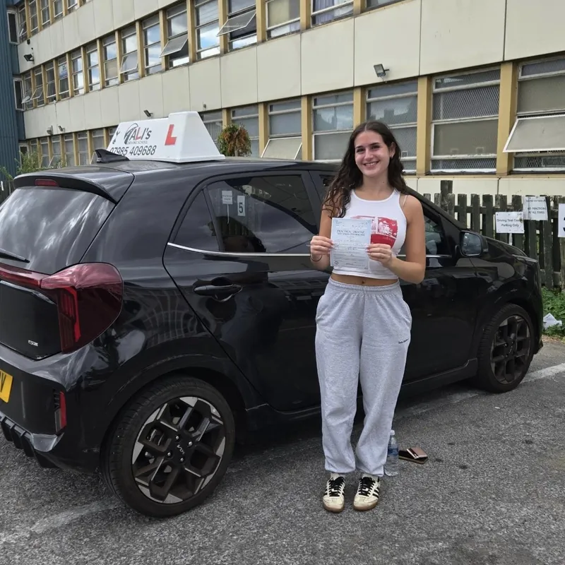 A new driver in a white top holding a pass certificate beside the learner car