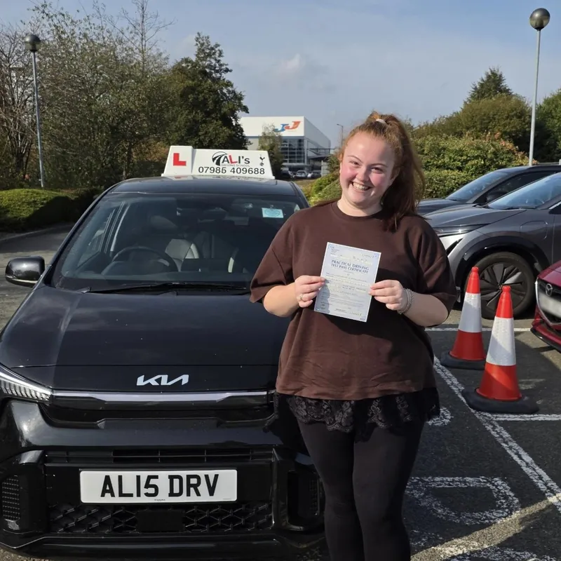 A new driver holding up a pass certificate beside the learner car, next to traffic cones